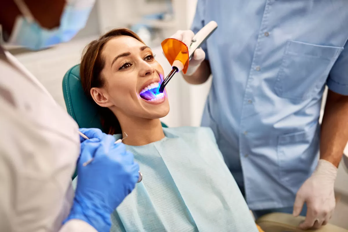 dentista e assistente alla poltrona con guanti di lattice agiscono con laser dentale luce blu su paziena donna seduta in poltrona